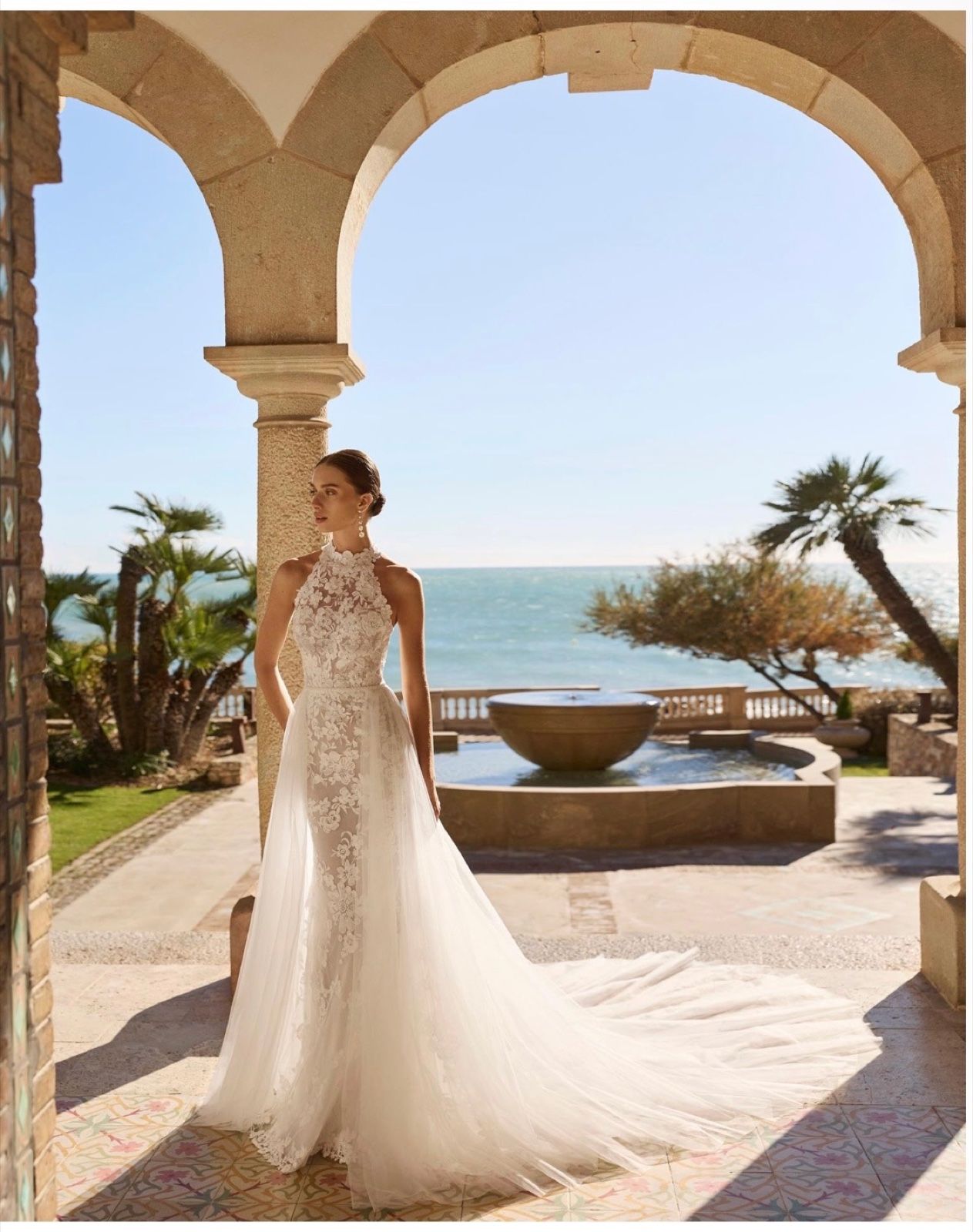 A halter-neck lace gown with a sweeping tulle overskirt, under a stone arch