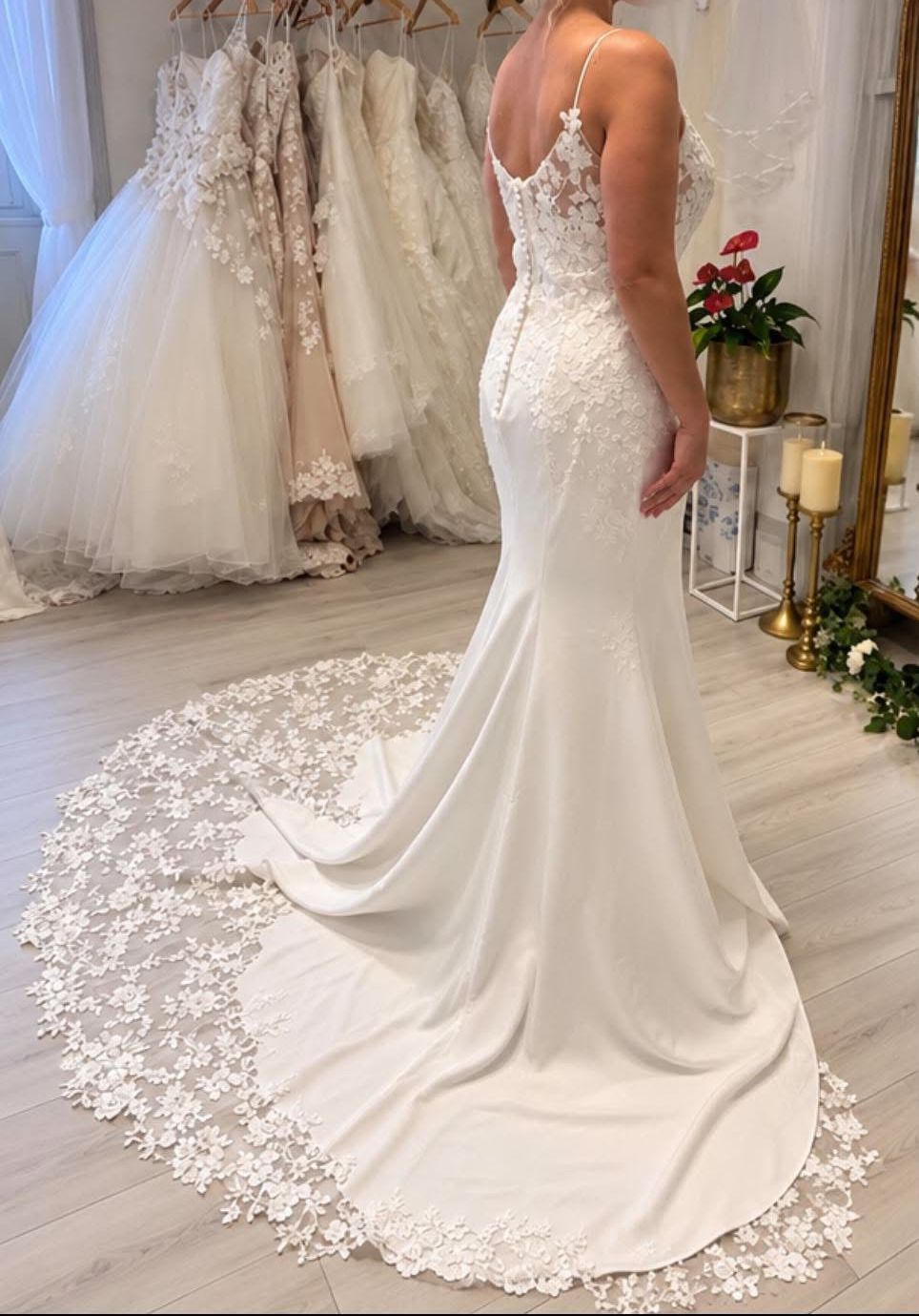 A fitted crepe gown with lace straps and a sweeping floral lace train, beside rails of gowns in the boutique